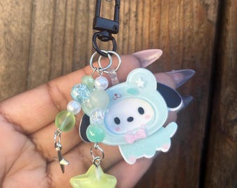 May include: A keychain featuring a cartoon character in a light blue outfit with a pink bow. The keychain has a black clasp, a star-shaped charm, and beads in shades of green and white. The background is a wooden surface.
