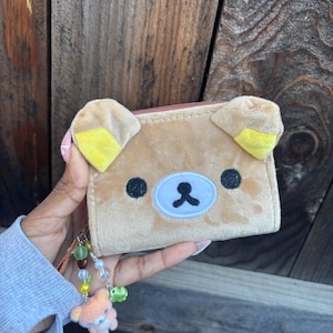May include: A tan plush wallet with a bear face design, featuring black eyes and a blue nose. The wallet has yellow-tipped ears and a beaded charm with a small bear. The background is a wooden surface.