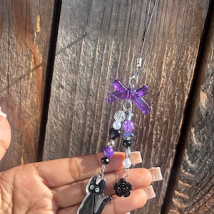 May include: A phone charm featuring a purple translucent bow at the top, two beaded strands with purple, black, and white beads, a black cat charm on one strand, and a black rose charm on the other, attached to a black cord with a metal clasp.