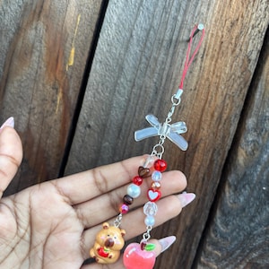 May include: A beaded charm with a clear bow, red string, and silver hardware. The charm features a small bear holding a red apple and a red apple charm. The beads are red, white, and clear.