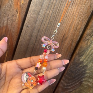 May include: A handmade charm featuring a cartoon character, a red star, and colorful beads. The charm has an orange cord and a clear bow. The cartoon character is orange and is next to a red and white pokeball.