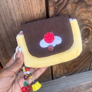 May include: A small yellow plush pouch with a brown corduroy flap featuring an embroidered red and white mushroom design. The pouch includes a metal keyring adorned with a beaded charm strand featuring a silver star, red cherries, and a yellow pudding.