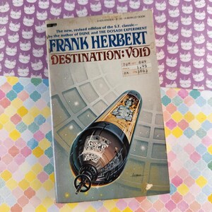 Puede incluir: Un libro de bolsillo vintage titulado "DESTINATION: VOID" de Frank Herbert. La portada presenta una nave espacial futurista con una figura en su interior. El libro está sobre un fondo de papel estampado con caras de gato y formas geométricas coloridas.