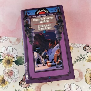 May include: A paperback book titled "Renunciates of Darkover" by Marion Zimmer Bradley. The cover has a purple border with illustrations of a snowy landscape, a person, and a white horse. The book is on a floral patterned surface.