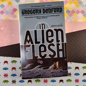 May include: A paperback book cover with a blue background and the title "In Alien Flesh" in white and black lettering. The author's name, Gregory Benford, is printed in white lettering. The cover features a black and white image of a lunar landscape with a spacecraft in the distance.