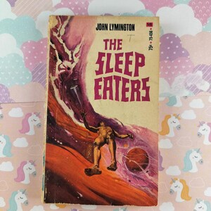 May include: Vintage paperback book titled "The Sleep Eaters" by John Lymington. The cover art features a figure falling through a red and purple space scene. The book is set against a pink background with unicorn and cloud patterns.
