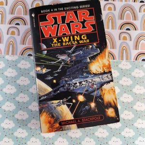 May include: A copy of "Star Wars: X-Wing The Bacta War" by Michael A. Stackpole. The book cover depicts a space battle with X-wing fighters and explosions. The book is part of the "Exciting Series!" and is set against a background of a rainbow pattern and a cloud pattern.