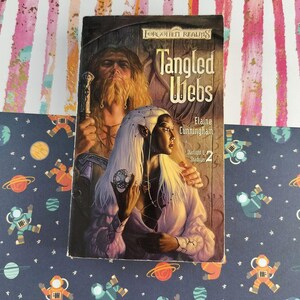 May include: A paperback book titled "Tangled Webs" by Elaine Cunningham, part of the Forgotten Realms series. The cover features a fantasy illustration of a woman with white hair and a man with a long beard. The background is a colorful pattern.