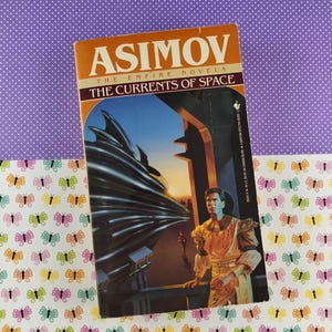 Könnte beinhalten: Ein Vintage-Taschenbuch mit dem Titel "The Currents of Space" von Isaac Asimov. Das Cover zeigt eine futuristische Illustration mit einem Mann in goldener Kleidung und einer dunklen, abstrakten Struktur. Das Buch liegt auf einem lila gepunkteten Hintergrund und einem Papier mit Schmetterlingsmuster.