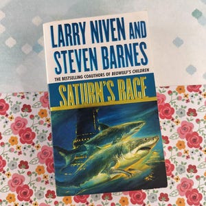May include: A paperback book titled "Saturn's Race" by Larry Niven and Steven Barnes. The cover features a dark blue underwater scene with two sharks. The title and author's names are in large, bold, blue letters.