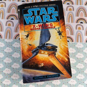 May include: A paperback book titled "Star Wars: X-Wing Wedge's Gamble" with a striking cover illustration. The cover shows a spacecraft in a fiery space scene, with the Star Wars logo. The book is part of a series, as indicated on the cover.