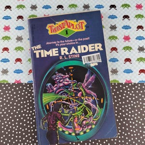 Peut inclure: Un livre de poche vintage intitulé "The Time Raider" de R.L. Stine, faisant partie de la série "Twist-A-Plot". La couverture présente une illustration colorée d'une scène futuriste. Le livre est posé sur un fond à motifs.