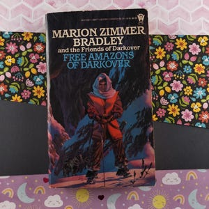 May include: A paperback book cover featuring a blue and white illustration of a person in a red and brown outfit standing in a snowy landscape. The book title is "Free Amazons of Darkover" and the author is "Marion Zimmer Bradley".