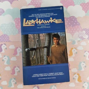 May include: A vintage paperback book titled "Ladyhawke" by Joan D. Vinge. The cover features a movie still with Matthew Broderick. The book is blue with gold lettering and includes the text "See the Spellbinding Fantasy Film."