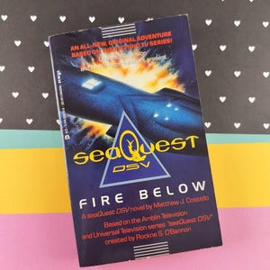 May include: A paperback book titled "SeaQuest Fire Below" based on the TV series. The cover features a blue submarine design with a yellow triangle logo and the text "SeaQuest DSV". The background has a fiery, underwater scene.