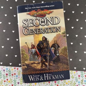 May include: A paperback book titled "The Second Generation" by Margaret Weis & Tracy Hickman. The cover features a fantasy illustration of three figures in armor and robes, with a castle in the background. The book is part of the "Dragonlance" series.