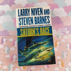 May include: A paperback book titled "Saturn's Race" by Larry Niven and Steven Barnes. The cover features a blue and yellow underwater scene with sharks. The background is a pink and white unicorn and cloud pattern.