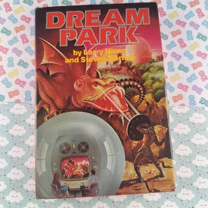 May include: A vintage paperback book titled "DREAM PARK" by Larry Niven and Steven Barnes. The cover art features a red dragon, a futuristic scene, and a warrior. The book is set against a background of a light blue and white patterned surface.