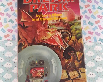 May include: A vintage paperback book titled "DREAM PARK" by Larry Niven and Steven Barnes. The cover art features a red dragon, a futuristic scene, and a warrior. The book is set against a background of a light blue and white patterned surface.
