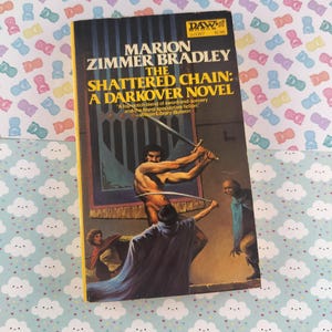 May include: A paperback book titled "The Shattered Chain: A Darkover Novel" by Marion Zimmer Bradley. The cover art shows a fantasy scene with figures wielding swords. The book has a yellow border and the DAW logo.