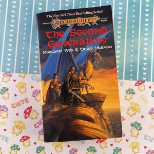 May include: A paperback book titled "The Second Generation" from the Dragonlance Saga series. The cover features a fantasy illustration of a warrior, a dragon, and another figure on a cliff, with a sunset backdrop. The book is on a patterned surface.