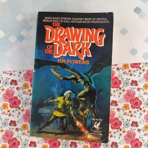 May include: A vintage paperback book titled "The Drawing of the Dark" by Tim Powers. The cover features a dark blue background with a winged demon and figures in yellow and brown. The book is a fantasy novel.