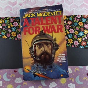 Puede incluir: Una novela de ciencia ficción de bolsillo titulada "A Talent for War" de Jack McDevitt. La portada presenta una nave espacial futurista con la cara de un hombre en la cabina.