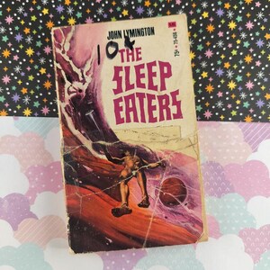 May include: Vintage paperback book titled "The Sleep Eaters" by John Lymington. The cover features a distressed illustration of a figure and a planet against a purple and orange background. The book is worn with visible signs of age.