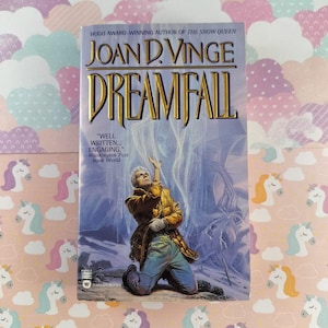 May include: A paperback book titled "DREAMFALL" by Joan D. Vinge. The cover features a man kneeling with his arm raised, with a blue and white background. The book has gold lettering and the words "WELL WRITTEN... ENGAGING." are printed on the cover.