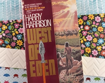 May include: A paperback book cover with a red background and gold lettering. The title is "West of Eden" by Harry Harrison. The cover art features a man and woman in a prehistoric setting with a spear in the foreground.