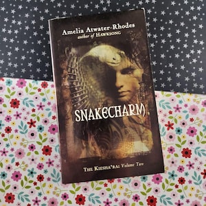 以下が含まれることがあります： Amelia Atwater-Rhodesの「Snakecharm」というタイトルのペーパーバック。シリーズ「The Kiesha'ra: Volume Two」の一部。表紙には、ヘビが重ねられた人物の肖像画と暗い背景が描かれています。本は花柄と星柄の表面に置かれています。