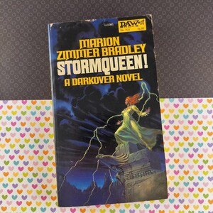 May include: Vintage paperback book titled "Stormqueen! A Darkover Novel" by Marion Zimmer Bradley. The cover art depicts a woman in a flowing green gown with red hair, standing on a stone structure, with lightning and a dark sky in the background.