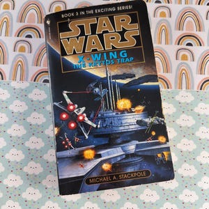 May include: A paperback book titled "Star Wars: X-Wing: The Krytos Trap" by Michael A. Stackpole. The cover features a space battle scene with X-wing fighters and a large space station. The book is set against a background of a rainbow pattern and a cloud pattern.