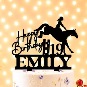 Könnte beinhalten: Eine weiße Torte mit Blumendekoration und einem schwarzen Tortenaufsatz mit Reiter und Pferd sowie dem Schriftzug "Happy Birthday #19 EMILY". Die Torte steht auf einem weißen Ständer, mit roten Blumen und einem warmen, verschwommenen Hintergrund.