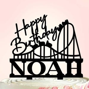 May include: Black cake topper with the words "Happy Birthday" and the name "Noah". The topper features a roller coaster design. The cake is decorated with pink and white frosting.