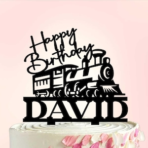 Op de afbeelding: Zwarte taarttopper met een treindesign en de woorden "Happy Birthday DAVID". De taart is versierd met witte frosting en roze en witte bloemblaadjes.