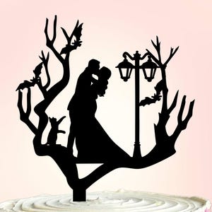 May include: Black silhouette cake topper depicting a kissing couple, bats, and a cat on a tree branch. A vintage street lamp is also included. The topper sits atop a white cake decorated with pink and white floral icing.