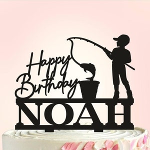 Op de afbeelding: Zwarte silhouet taarttopper van een jongen die vist met de tekst "Happy Birthday NOAH" op een witte taart.