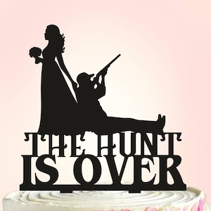 May include: Black silhouette cake topper of a bride and groom. The groom is holding a rifle and the bride is holding a bouquet. The text "The Hunt Is Over" is below the figures.
