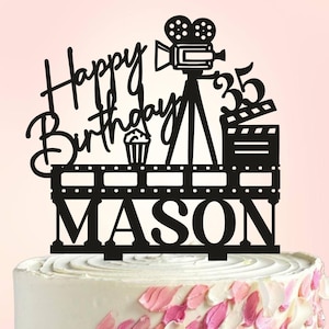 May include: A black cake topper with the words "Happy Birthday" and "Mason" in a script font. The topper features a film camera, a clapperboard, a cupcake, and the number 35. The cake has white frosting and pink and white petal decorations.