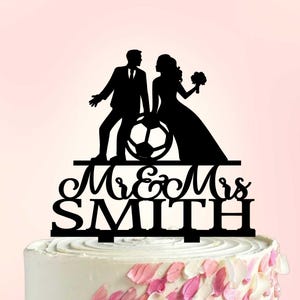 May include: Black silhouette cake topper featuring a bride and groom with a soccer ball. The topper reads "Mr & Mrs Smith" in elegant script. The cake is decorated with pink and white frosting.