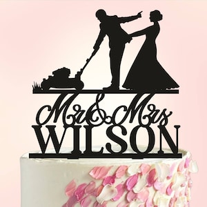 Pode incluir: Topper de bolo em silhueta preta de uma noiva e um noivo. O noivo está empurrando um cortador de grama e a noiva está segurando o braço dele. O texto "Mr. & Mrs. WILSON" está abaixo das figuras.