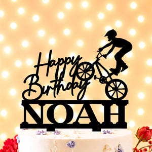 Puede incluir: Adorno negro para pastel con las palabras "Happy Birthday NOAH" y la silueta de una persona montando una bicicleta BMX. El adorno está colocado sobre un pastel blanco con decoraciones florales moradas y doradas.