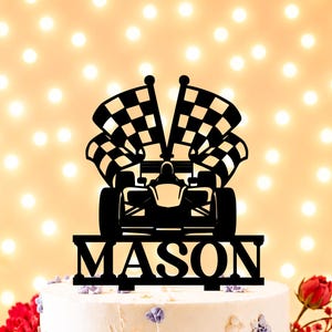 Peut inclure: Un cake topper noir représentant une voiture de course avec deux drapeaux à damiers. Le nom "MASON" est affiché en lettres grasses sous la voiture. Le topper est placé sur un gâteau blanc avec des décorations florales.