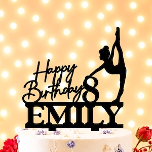 Peut inclure: Décoration de gâteau noire avec l'inscription "Happy Birthday 8 Emily" et la silhouette d'une gymnaste. Le gâteau blanc est orné de fleurs violettes et de détails dorés, parfait pour un anniversaire.