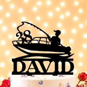 Puede incluir: Un adorno para pastel negro que presenta la silueta de una persona en un bote sosteniendo una caña de pescar, con el número 38 integrado en el diseño y el nombre DAVID en letras mayúsculas negritas en la base, colocado sobre un pastel blanco.
