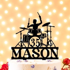 May include: Black cake topper depicting a drummer playing a drum kit. The topper includes the name "MASON" and the number "35". The cake is decorated with purple and gold floral accents and red roses.