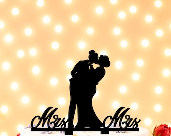 May include: A black silhouette cake topper featuring two figures kissing, with the word Mrs on the left and the word Mrs on the right, positioned on top of a white cake decorated with small floral accents and surrounded by red flowers.