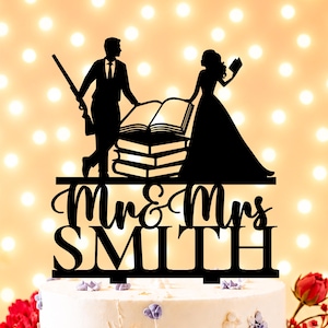 Puede incluir: Un adorno para pastel en silueta negra que muestra a una persona con traje sosteniendo un rifle y a otra con vestido sosteniendo un libro, junto a una pila de libros. El texto "Mr & Mrs SMITH" aparece en fuente cursiva y de bloque.