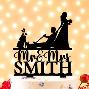 Puede incluir: Un adorno para pastel en silueta negra que muestra a una persona sentada con un palo y bolsa de golf, y otra de pie sosteniendo un ramo. El texto Mr & Mrs SMITH aparece en una combinación de letra cursiva y de molde en la base.